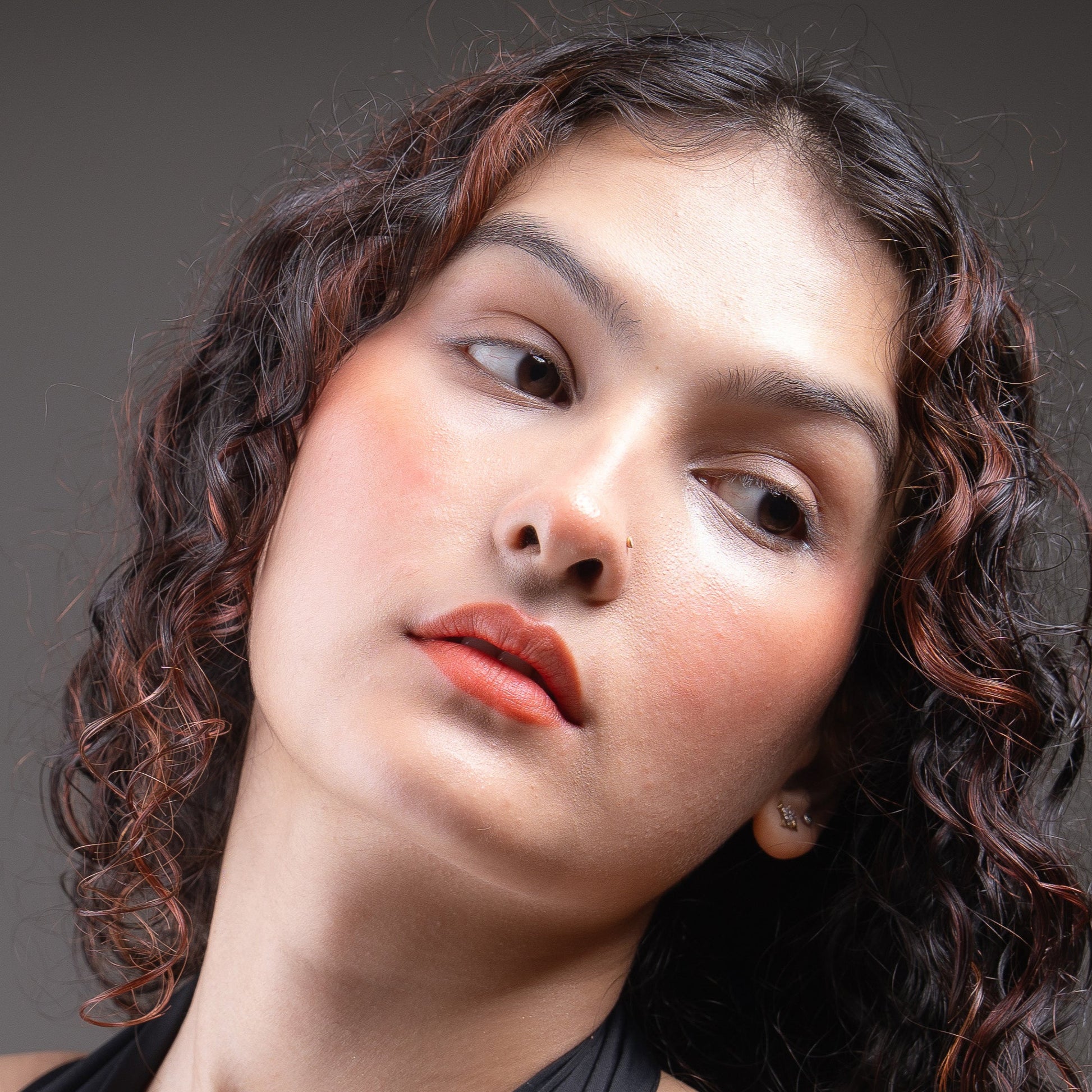 Woman wearing a black sleeveless top against a gray background wearing warm rust coral colour lip and cheek tint
