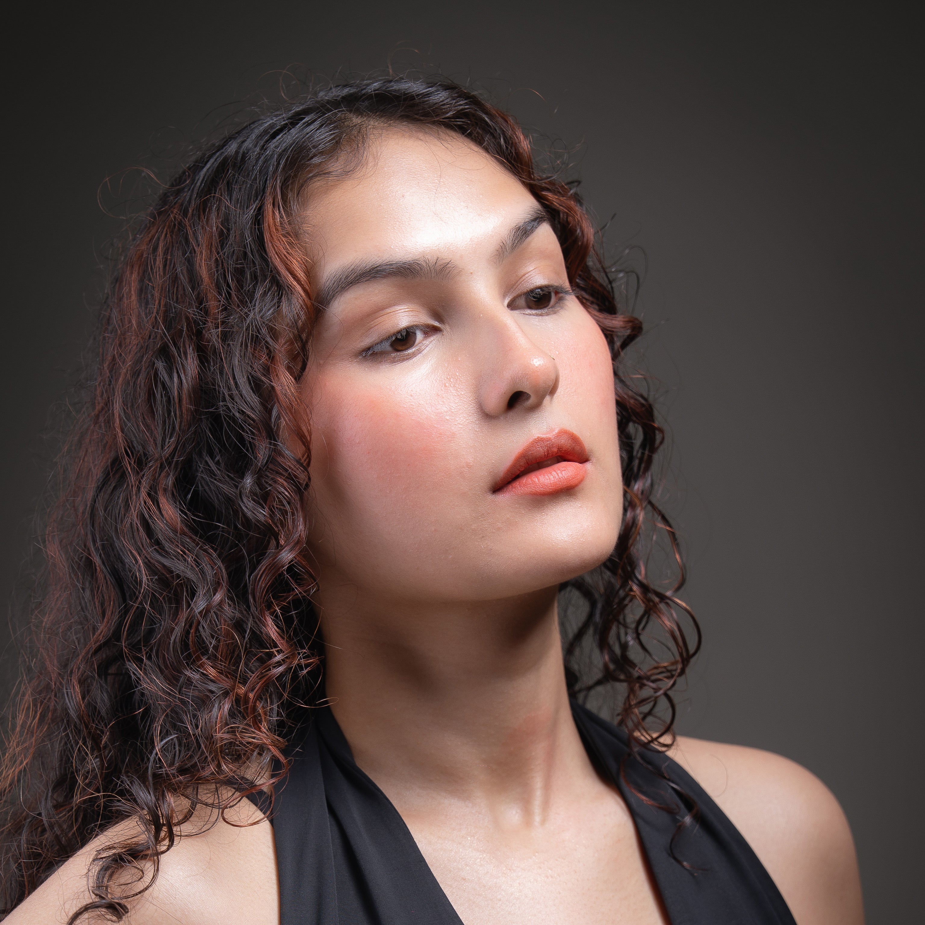 Woman with styled hair posing against a gray background wearing a sunset red lip and cheek tint